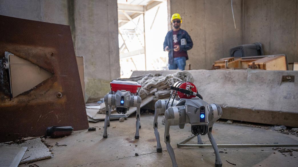 A man wearing a hard hat in the background oversees a pair of four-legged robots as they navigate through a structure in a demonstration of their abilities.